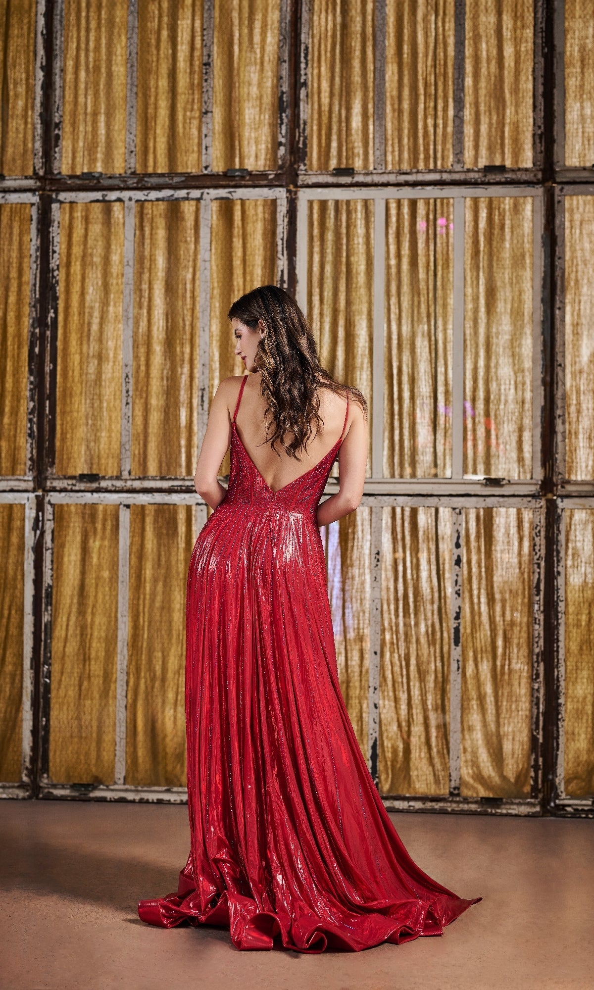 A woman wearing the Long Prom Dress: Tiffany 16189, a metallic pleated gown, stands with her back to the camera, facing gold curtains and a window framed by a rustic wooden grid.